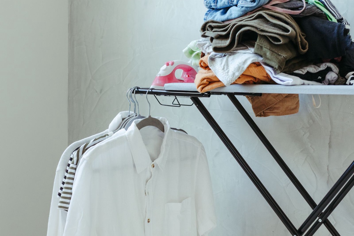 Folded laundry stacked on an ironing board above a white shirt on hangers in a laundry room
